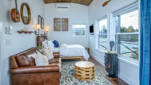 Una habitación acogedora con un sofá de cuero marrón, una cama con ropa de cama blanca, un taburete de madera redondo y una estufa de leña.