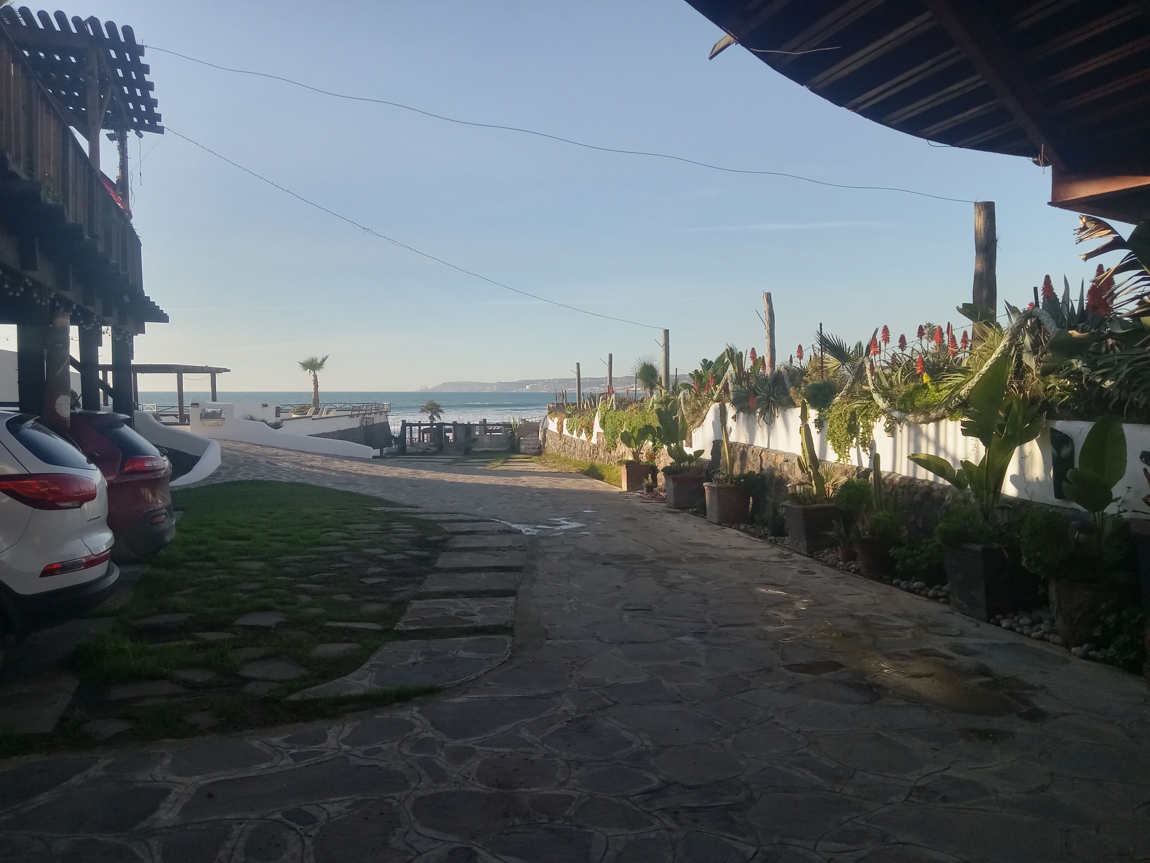 Un sendero pavimentado que conduce a una playa, con coches aparcados y una pared blanca con plantas en macetas a la derecha.