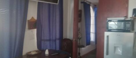 Um quarto com uma mesa de jantar, cadeiras, uma televisão na parede e uma geladeira com micro-ondas em cima.