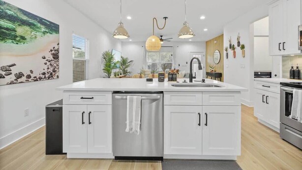 Una cocina moderna con armarios blancos, una isla central, lavavajillas, fregadero y estufa. Hay luces colgantes, un ventilador de techo y un ventanal que proporciona luz natural.
