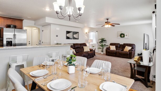 A modern kitchen with a refrigerator, dining table set for six, and a living area with a sofa and a ceiling fan.