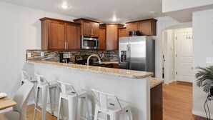 A modern kitchen with wooden cabinets, stainless steel appliances, a granite countertop, and a bar area with white stools.