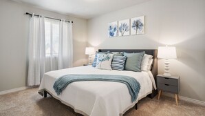 A bedroom with a bed, two bedside tables with lamps, a window with curtains, and wall art.