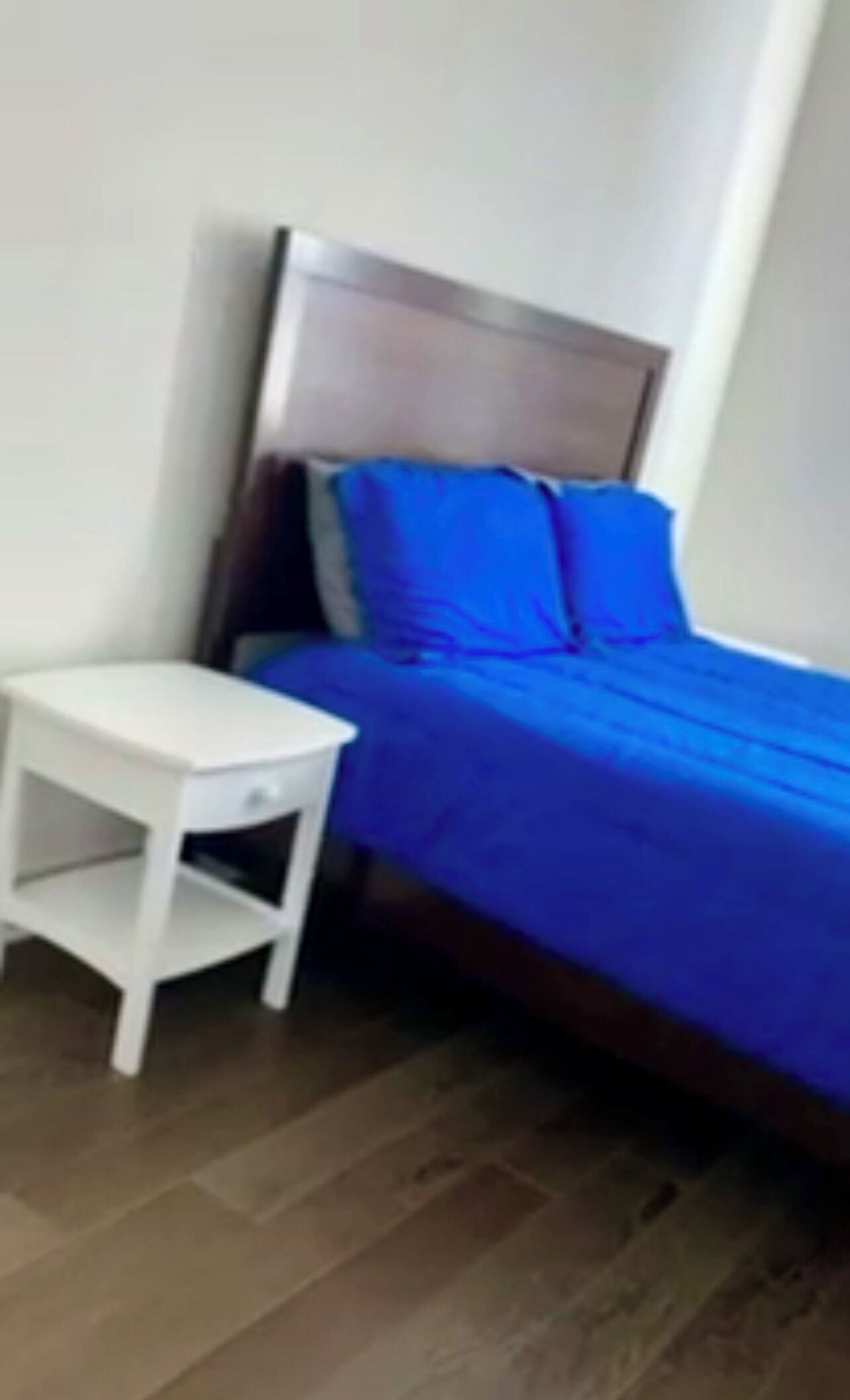 Een slaapkamer met een bed, een klein wit nachtkastje en een houten vloer.