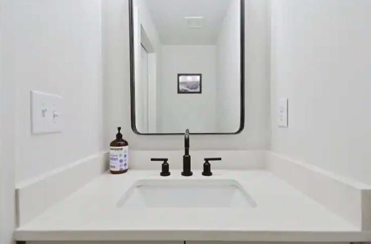 A white bathroom with a rectangular sink, a black faucet, and a mirror above it. There is a bottle of hand soap on the left side of the sink.