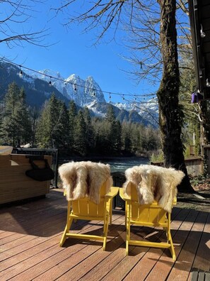 Zwei gelbe Stühle mit Felldecken auf einer Holzterrasse mit Blick auf einen Fluss und Berge.