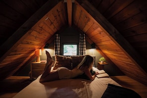 Ein gemütliches Schlafzimmer im Dachgeschoss mit geneigten Holzbalken, einem Bett mit Kissen, einem Nachttisch mit Lampe und einem Fenster mit Blick.