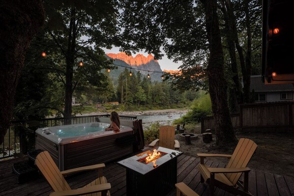 A hot tub with a fire feature, surrounded by wooden deck chairs, overlooking a river and mountains.