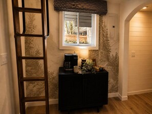 Ein Zimmer mit einer Holzlade, einem Fenster mit einem gemusterten Vorhang, einer Kaffeemaschine, einer Tasse und einer kleinen Pflanze auf einem dunklen Schrank.