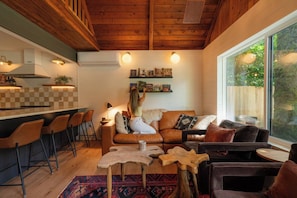 Ein gemütliches Wohnzimmer mit Holzbalkendecke, einem großen Fenster, einer Küchenzeile mit Barhocker und einem Couchtisch.
