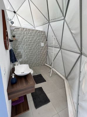 A modern bathroom with a geometric patterned wall, a wooden vanity, a white sink, and a shower area with a white canopy. - Domo Geodésico - Conforto e Relaxamento na Natureza (Socorro)