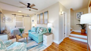 A living room with a teal sofa, a coffee table, a side table with a lamp, a ceiling fan, and a staircase with a plant. - Newly remodeled house in San Marco few minutes walk to Square  & Riverwalk (Jacksonville)