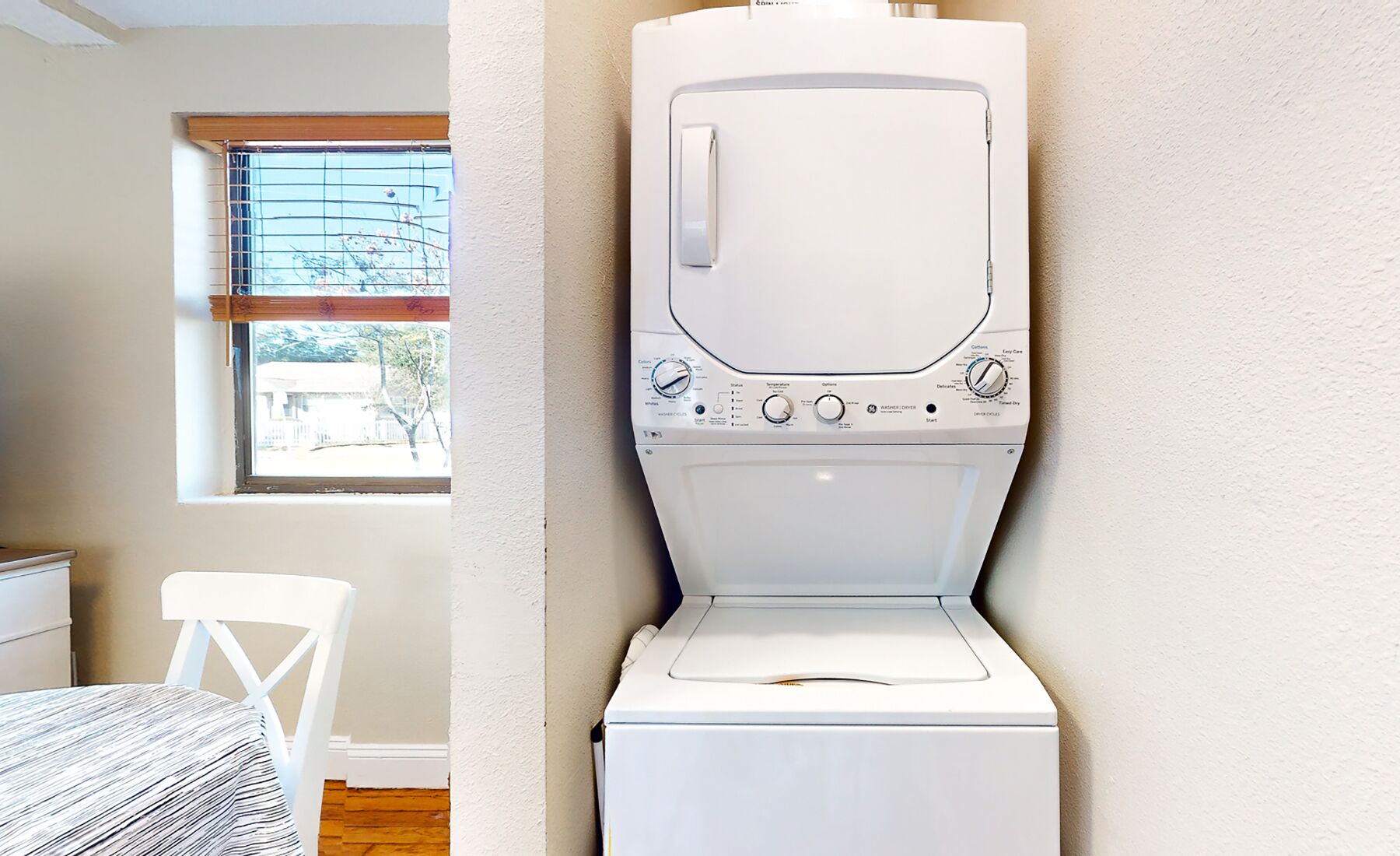 A compact, white laundry room with a front-loading washer and dryer. A window with blinds is visible, and a white chair is placed near the window.
