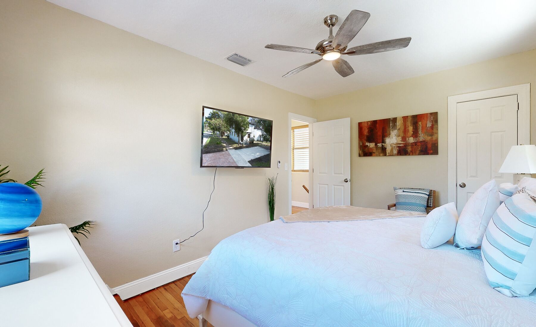 A bedroom with a bed, a ceiling fan, a TV mounted on the wall, and a door leading to another room.