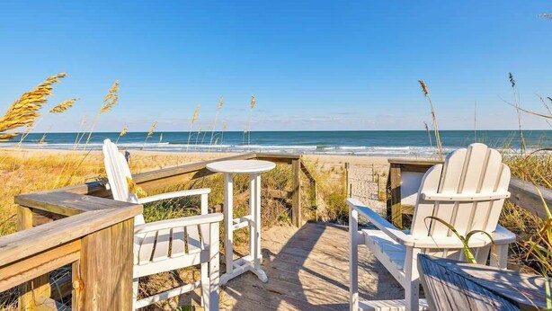 A wooden deck with two white Adirondack chairs and a small round table, overlooking a beach with the ocean in the background. - A Shore Thing - Oceanfront - Hot Tub -Pet Friendly (Carolina Beach)