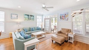 Un salon spacieux avec un canapé bleu clair, un fauteuil beige et une table basse blanche. Deux tableaux ornent le mur et un ventilateur de plafond est installé au centre.