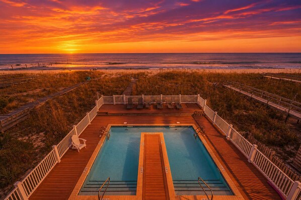 Una piscina con cubierta de madera, rodeada por una valla de madera, con vistas a una playa al atardecer.