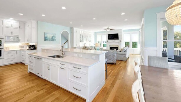 A spacious kitchen with white cabinetry, a central island, and a dining area with a wooden table and chairs. - Seaside Palace (Kure Beach)