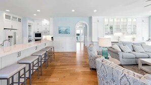 A modern kitchen with white cabinetry, a central island with stools, and a dining area with a grey sofa and patterned armchairs. - Seaside Palace (Kure Beach)