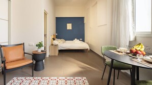 Ein kleines Zimmer mit Bett, Stuhl, einem kleinen Tisch und einer Obstschale, sowie einem Fenster mit Vorhängen.