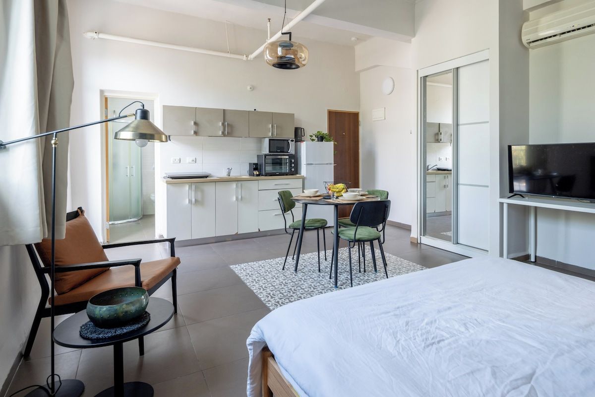 A modern hotel room with a bed, a desk, a chair, a dining table with chairs, a kitchen area, and a television.