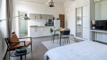A modern hotel room with a bed, a desk, a chair, a dining table with chairs, a kitchen area, and a television.