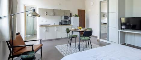 A modern hotel room with a bed, a desk, a chair, a dining table with chairs, a kitchen area, and a television.