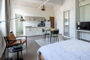 A modern hotel room with a bed, a desk, a chair, a dining table with chairs, a kitchen area, and a television.