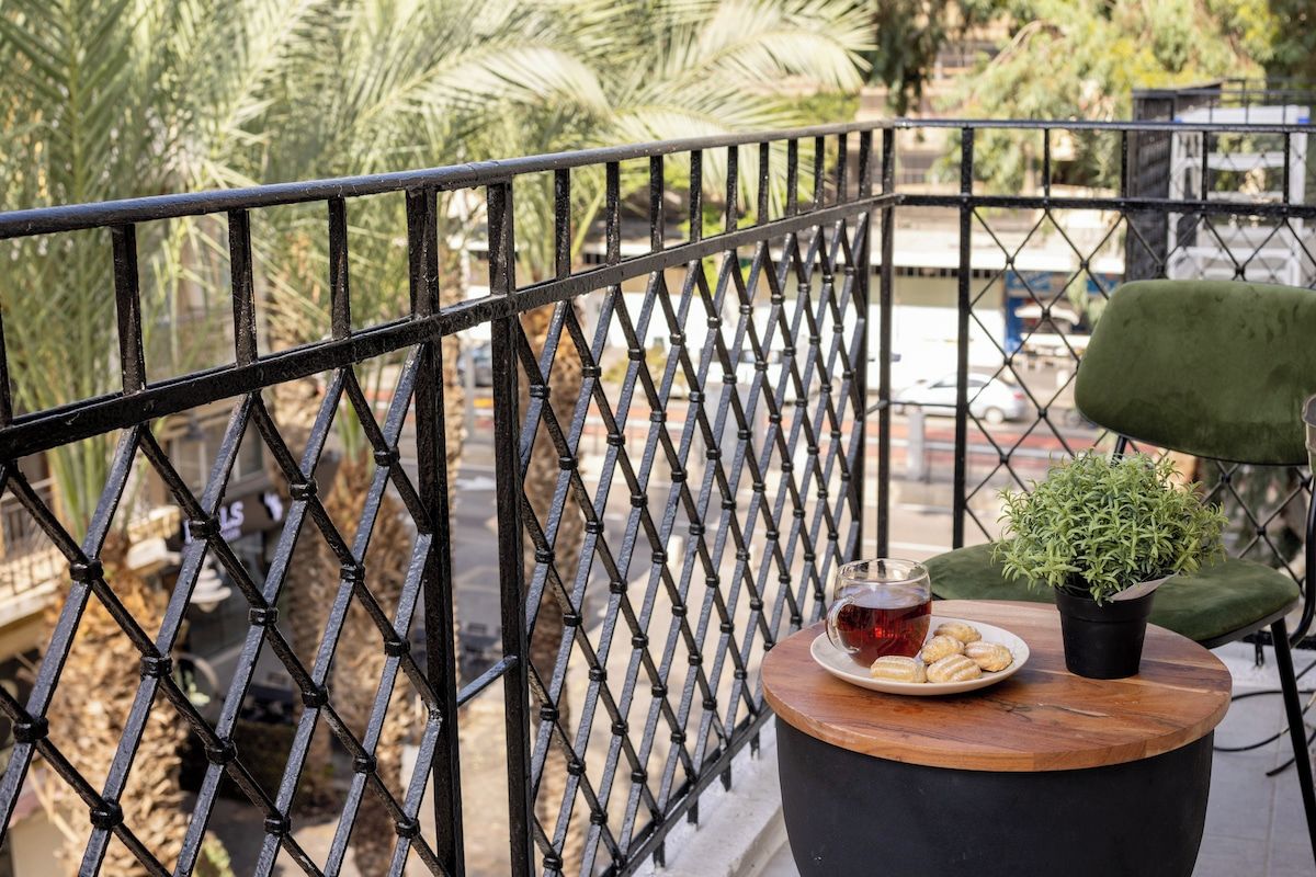 A balcony with a metal railing, a green chair, a small table with a plate of cookies, a glass of red drink, and a potted plant.