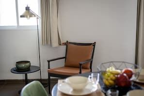 Une pièce comprenant une chaise, une petite table avec un bol, une table à manger avec des assiettes et un panier de fruits.