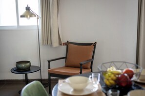 A room with a chair, a small table, a bowl, a fruit bowl, and a lamp.