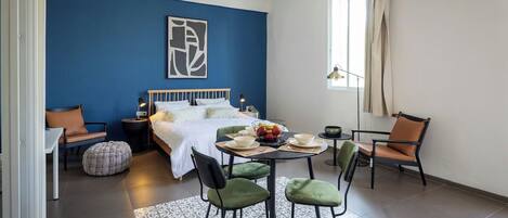 A modern bedroom with a blue accent wall, a bed with white linens, a dining table with chairs, and a window with curtains.