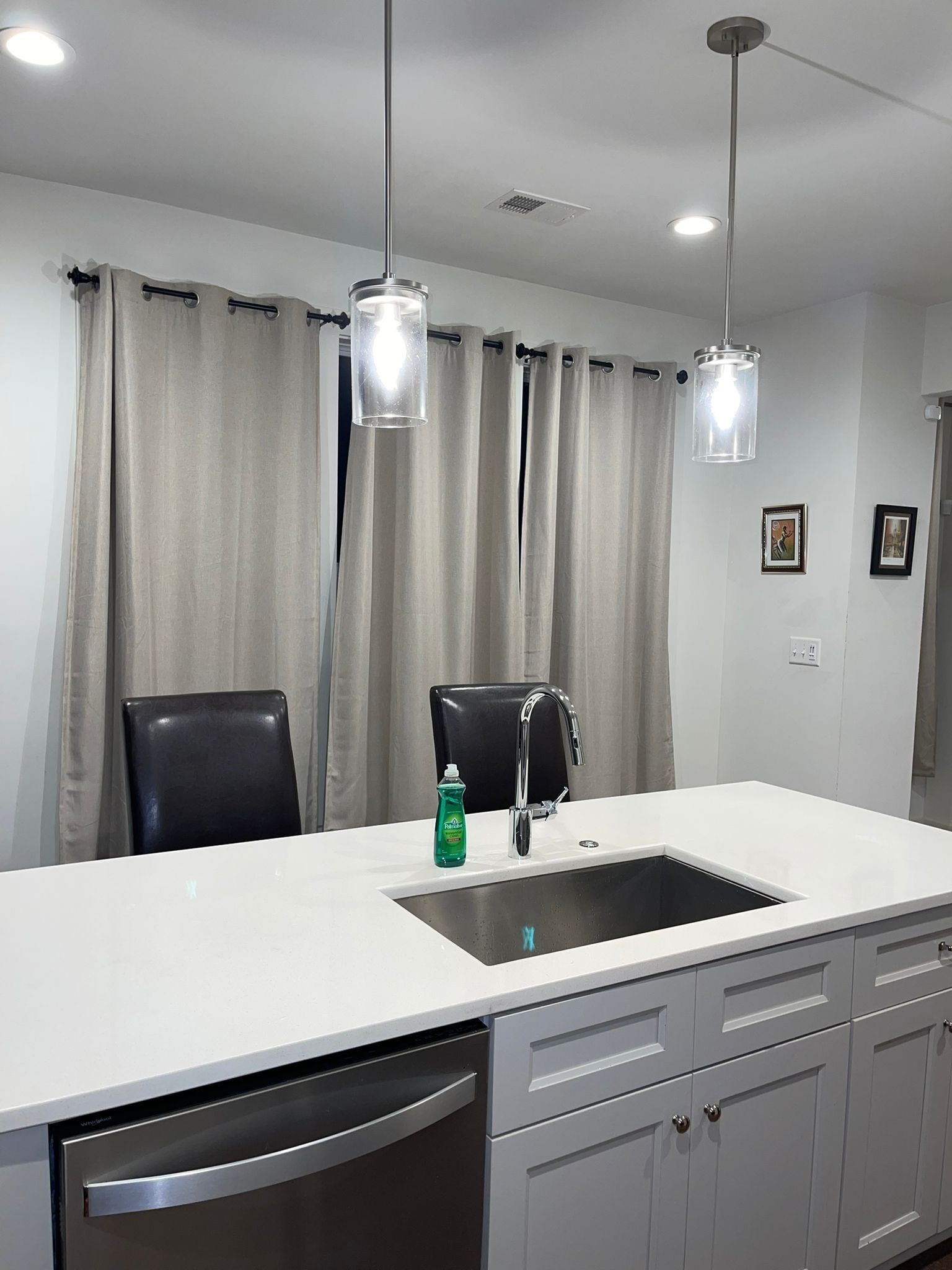 A modern kitchen with a white countertop, stainless steel sink, and black chairs. There are two pendant lights hanging above the sink.