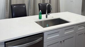 A modern kitchen with a white countertop, stainless steel sink, and black chairs. There are two pendant lights hanging above the sink.