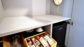 A kitchenette with a white countertop, a drawer with food items, a hanging garment, and a black appliance.