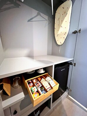 A kitchenette with a white countertop, a drawer with food items, a hanging garment, and a black appliance.