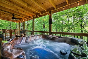 A hot tub with a wooden deck and chairs, surrounded by trees.