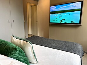 A bedroom with a bed, a television mounted on the wall displaying a tropical scene, and a door leading to another room.