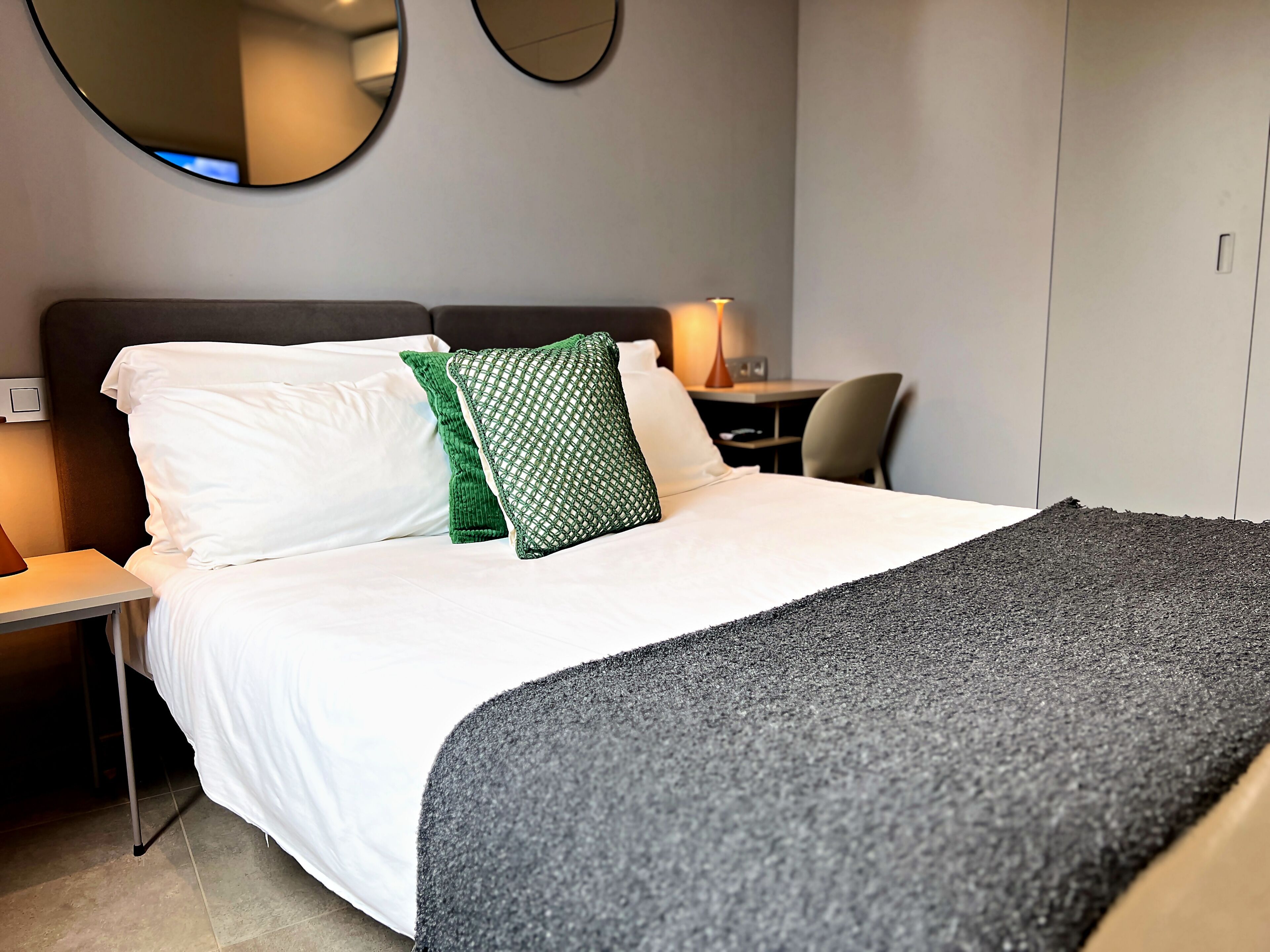 A hotel room with a bed, two pillows, a bedside table with a lamp, a chair, and a wardrobe.