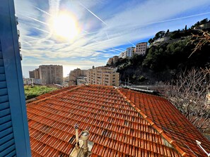 Um terraço com telhado de cerâmica tradicional, céu claro com rastros de aviões, e edifícios ao fundo.