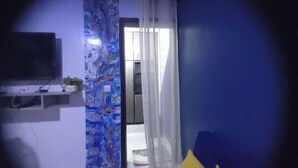 Una habitación con una pared azul, un televisor montado en la pared, una ventana con cortinas traslúcidas y una puerta que da a otra habitación.
