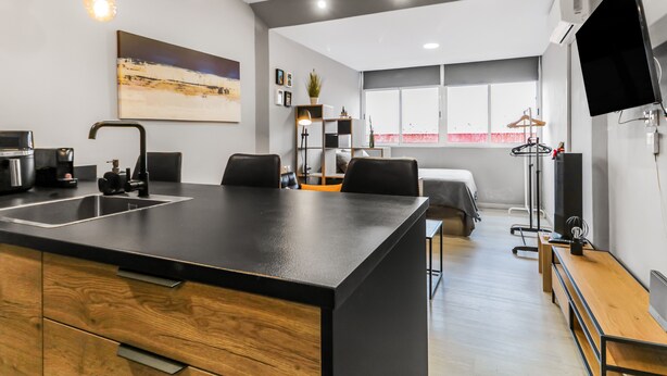A modern kitchen with a central island, a built-in sink, and a dining area with a bench. There is a large window with a view of the city, and a flat-screen TV mounted on the wall.