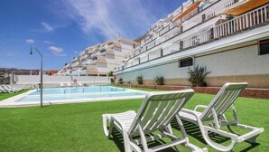 A pool area with two white lounge chairs, a swimming pool, and a building with balconies in the background.