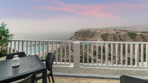 A balcony with a table and chairs, overlooking a coastal landscape with buildings and cliffs.
