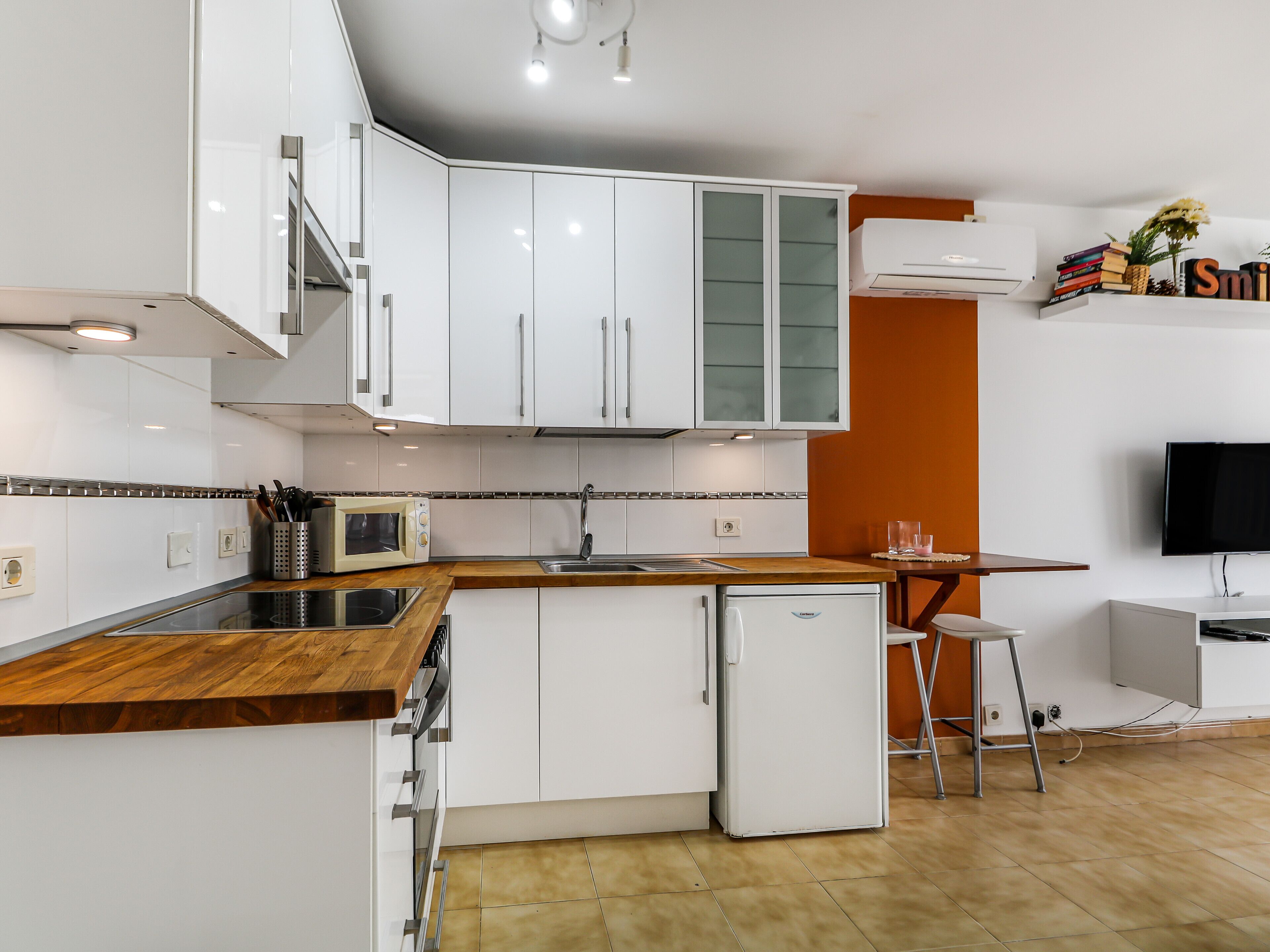 Une cuisine moderne avec des armoires blanches, un four à micro-ondes encastré, un évier et un petit réfrigérateur. On y trouve un plan de travail en bois et un coin repas avec une chaise.