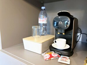 A coffee machine with a cup and saucer, a water bottle, and two packets of snacks on a table.