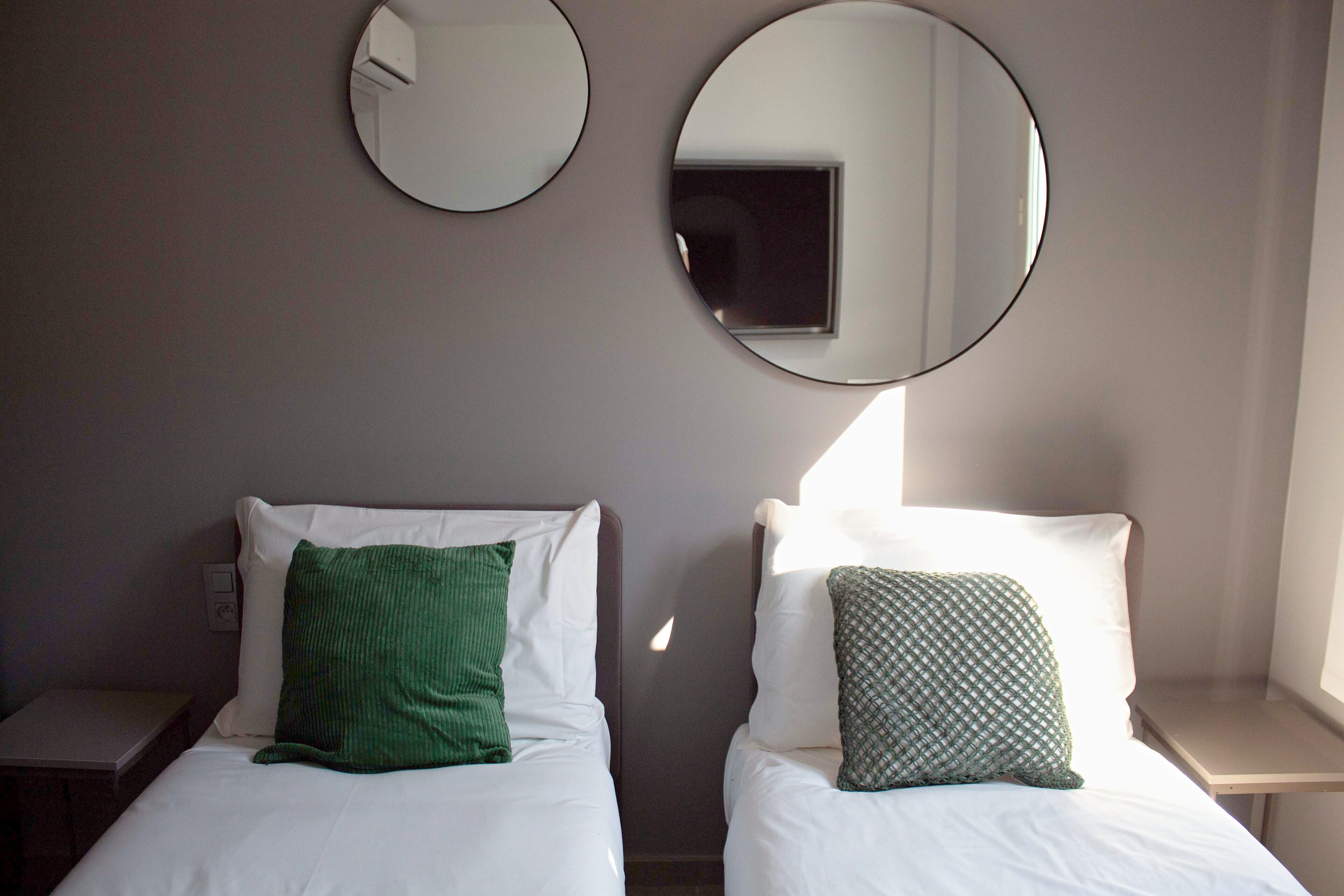 Two beds with white linens, green pillows, and two round mirrors on the wall.