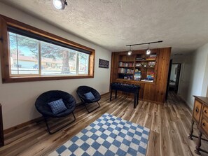 Una stanza con pavimento in legno, un tappeto bianco e blu a scacchi, due poltrone nere, un tavolo da calcio, una libreria e una finestra con vista su una casa e un albero.