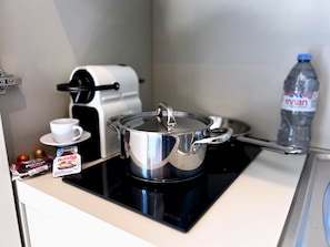 A kitchenette with a pot on an induction cooktop, a coffee maker, a cup, a Nutella packet, and a bottle of Evian water.