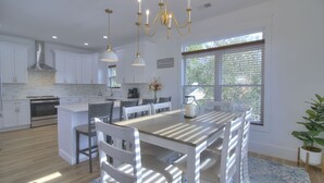 A modern kitchen with white cabinetry, a central island, pendant lighting, and a large window with blinds. - Inlet Escape - 5 BR/4 BA House Minutes From Murrells Inlet Marshwalk (Murrells Inlet)